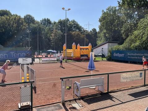 Tennisplatz mit Spielern, einem Sprungschloss im Hintergrund und grüner Landschaft unter blauem Himmel.