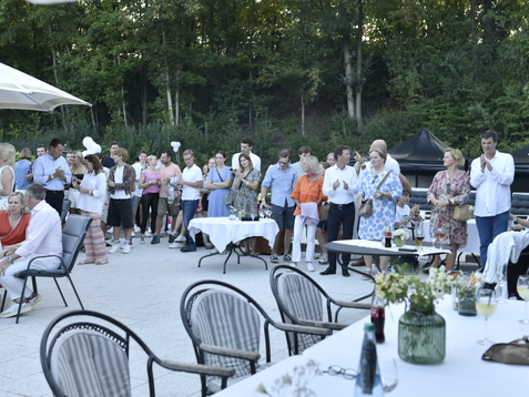 Gruppierung von Personen in einem Garten, applaudierend, mit Tischen und St&uuml;hlen im Vordergrund. B&auml;ume im Hintergrund sichtbar.