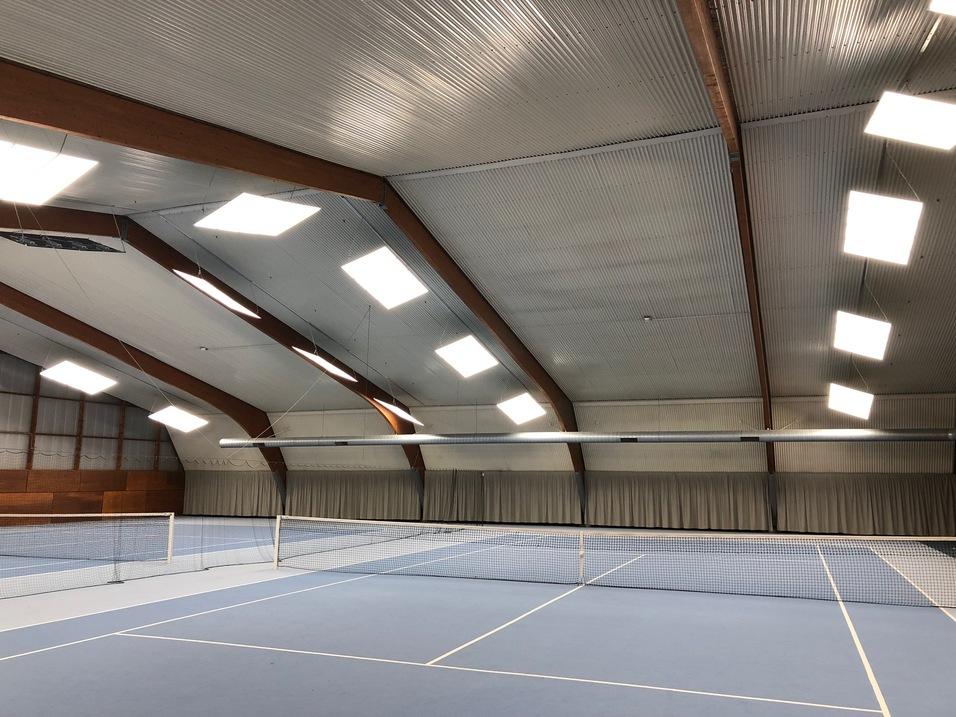 Blick in eine Tennis-Halle mit blauem Boden, Netzen und großen Dachfenstern.