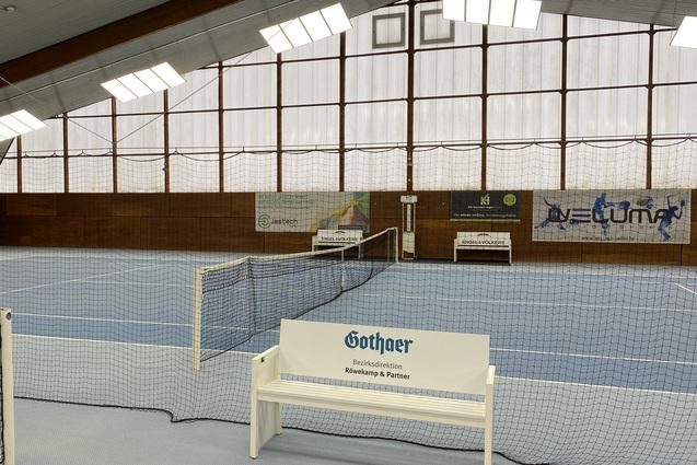 Innenansicht einer Tennishalle mit blauem Boden, Netzen und B&auml;nken an den Seiten. Gro&szlig;er Fensterbereich im Hintergrund.