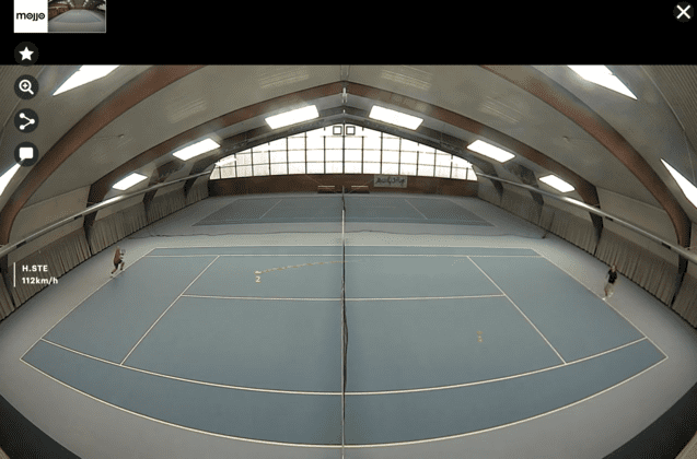 Innenansicht eines Tennisplatzes mit zwei Spielern, blauen Belag und Sicht auf das Hallendach und Fensterausschnitt.