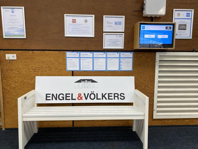 Wei&szlig;er Informationsstand mit der Aufschrift &bdquo;ENGEL & V&Ouml;LKERS&ldquo; und mehreren Zertifikaten an der Wand.