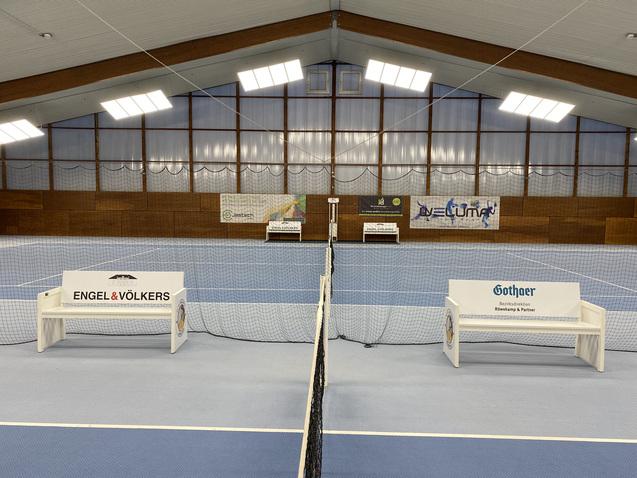 Blick in eine Tennishalle mit zwei B&auml;nken und Netzen auf einem blauen Platz, umgeben von Werbebannern.