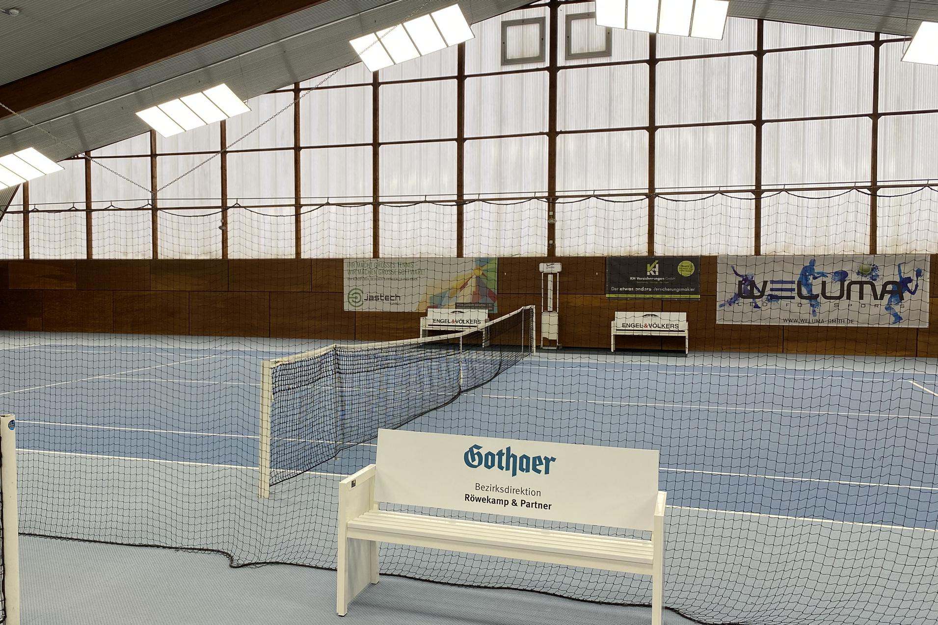 Innenansicht einer Tennishalle mit blauem Boden, Netzen und B&auml;nken an den Seiten. Gro&szlig;er Fensterbereich im Hintergrund.