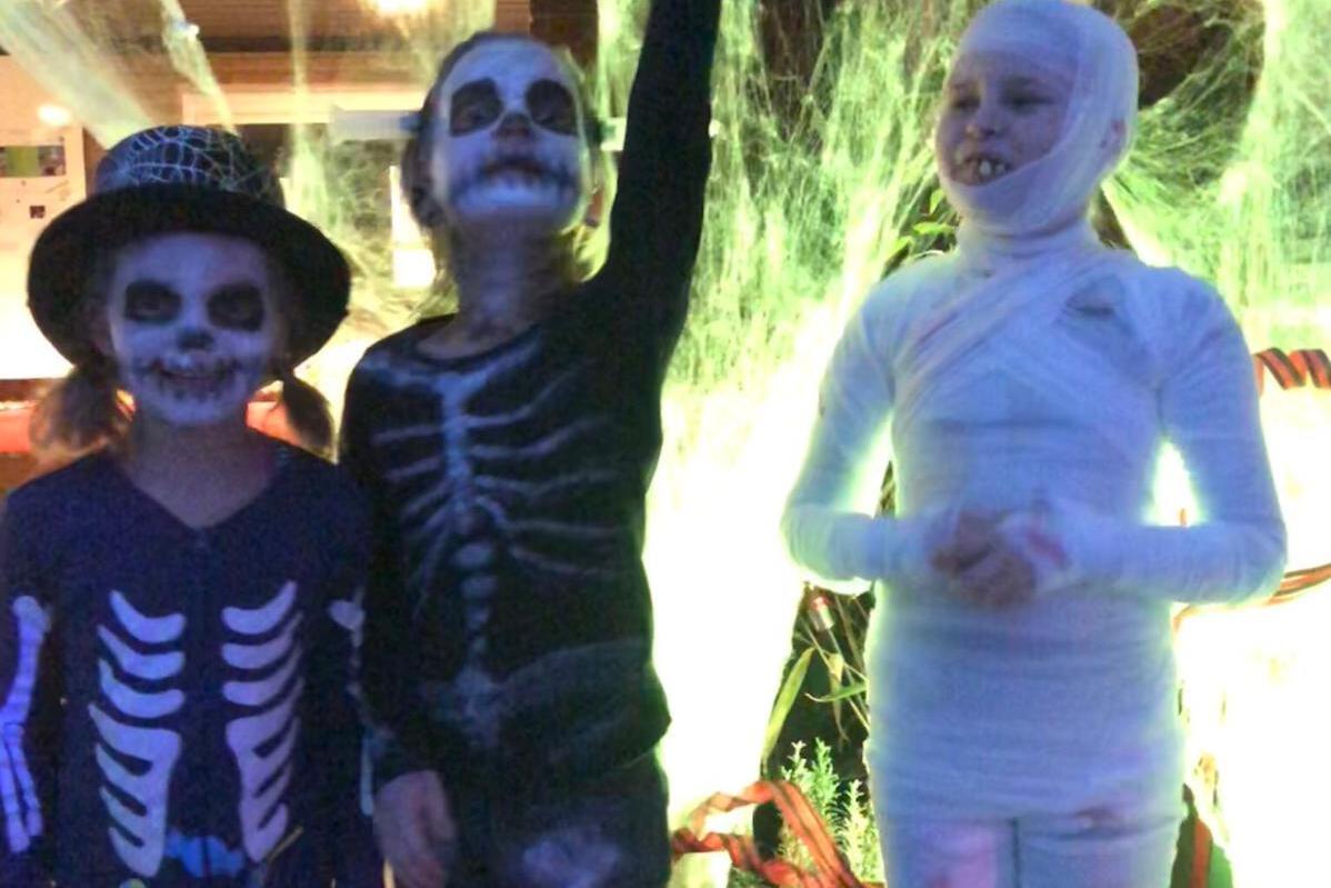 Drei Kinder in Halloween-Kost&uuml;men: zwei als Skelette, eines als Mumie, vor einer gruseligen Dekoration aus Spinnweben.