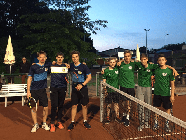 Gruppe von sieben Jugendlichen in Sportkleidung, l&auml;chelnd auf einem Tennisplatz bei Abendd&auml;mmerung, einige mit Schl&auml;gern.