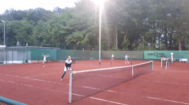 Zwei Tennisspielerinnen in Aktion auf einem Tennisplatz mit roten Sandboden, umgeben von B&auml;umen bei Abendlicht.