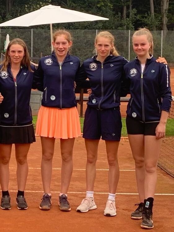 Vier junge Tennisspielerinnen in blauen Sportjacken stehen l&auml;chelnd auf einem Tennisplatz mit Sandbelag.
