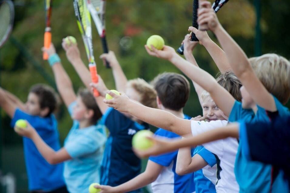 Gruppe von Kindern, die Tennisb&auml;lle hochwerfen, w&auml;hrend sie Schl&auml;ger in der Hand halten, auf einem Tennisplatz.