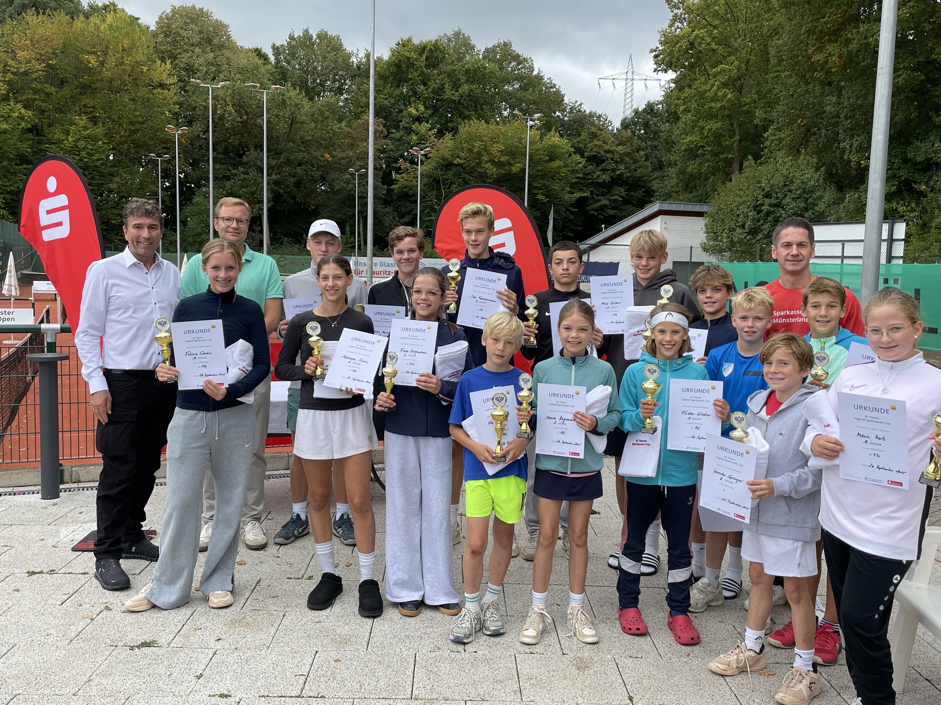 Gruppenfoto von jungen Tennisspielern mit Pokalen und Urkunden auf einem Tennisplatz, umgeben von Trainern und Sponsoren.