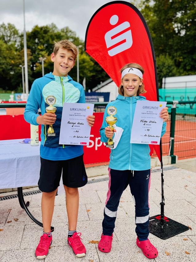 Zwei Kinder stehen mit Pokalen und Urkunden auf einem Sportplatz, im Hintergrund ein Banner und Tennispl&auml;tze.