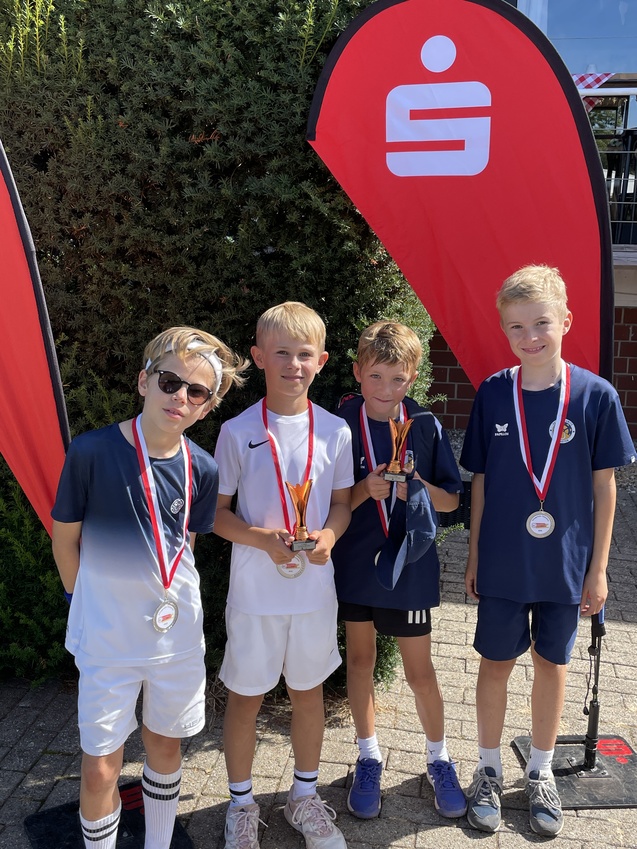 Vier Kinder in Sportkleidung stehen vor einem gr&uuml;nen Hintergrund, halten Pokale und Medaillen in der Hand.