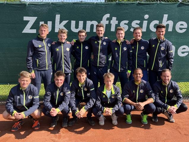 Gruppe von 14 jungen Tennis-Spielern in Sportjacken, l&auml;chelnd vor einem gr&uuml;nen Netz auf einem Tennisplatz.