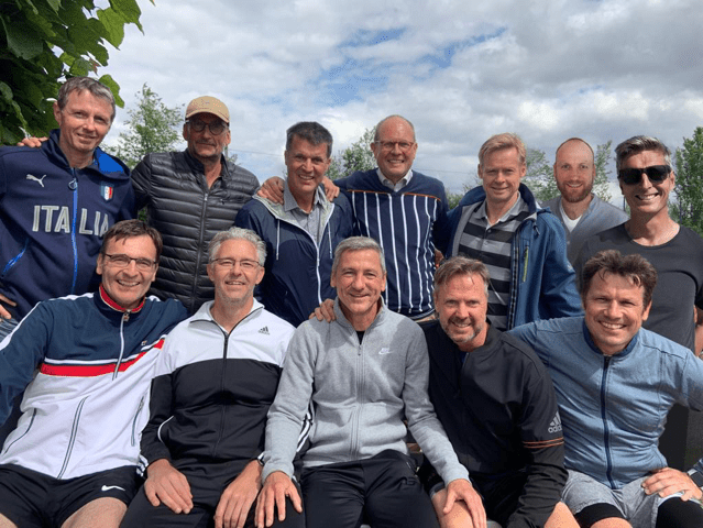 Gruppennahaufnahme von 12 M&auml;nnern, l&auml;chelnd, im Freien vor bew&ouml;lktem Himmel, in Sportkleidung.