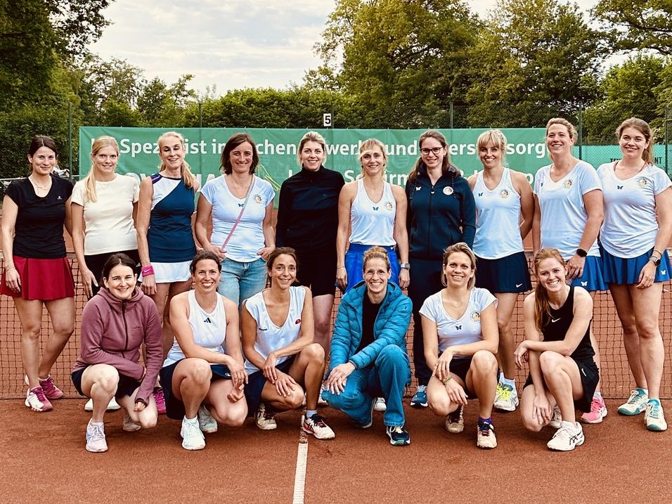 Gruppe von 16 Frauen in Sportkleidung posiert auf einem Tennisplatz mit B&auml;umen im Hintergrund.