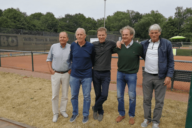 F&uuml;nf M&auml;nner stehen l&auml;chelnd nebeneinander auf einem Tennisplatz, im Hintergrund sind B&auml;ume und eine Tennisnetz zu sehen.