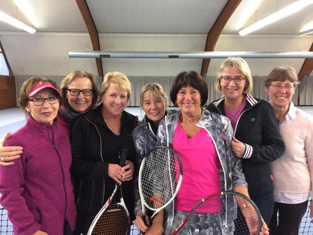 Gruppe von sieben Frauen in Sportkleidung mit Tennisschl&auml;gern, l&auml;chelnd in einer Tennishalle posierend.