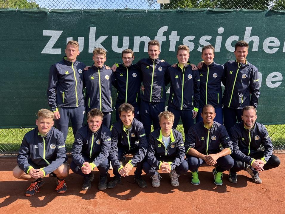 Gruppe von 14 jungen Tennis-Spielern in Sportjacken, l&auml;chelnd vor einem gr&uuml;nen Netz auf einem Tennisplatz.