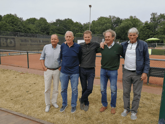 Fünf Männer stehen lächelnd nebeneinander auf einem Tennisplatz, im Hintergrund sind Bäume und eine Tennisnetz zu sehen.
