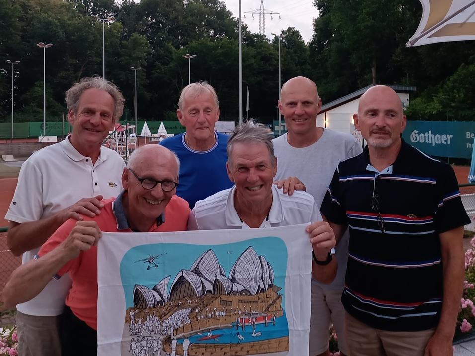 Sechs M&auml;nner posieren l&auml;chelnd auf einem Tennisplatz, halten ein buntes Bild mit bergigem Motiv in der Mitte.