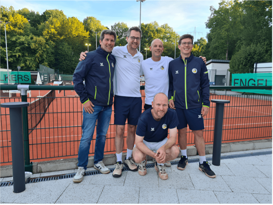 F&uuml;nf M&auml;nner posieren l&auml;chelnd vor einer Tennisanlage mit orangefarbenem Belag und gr&uuml;ner Umz&auml;unung im Hintergrund.