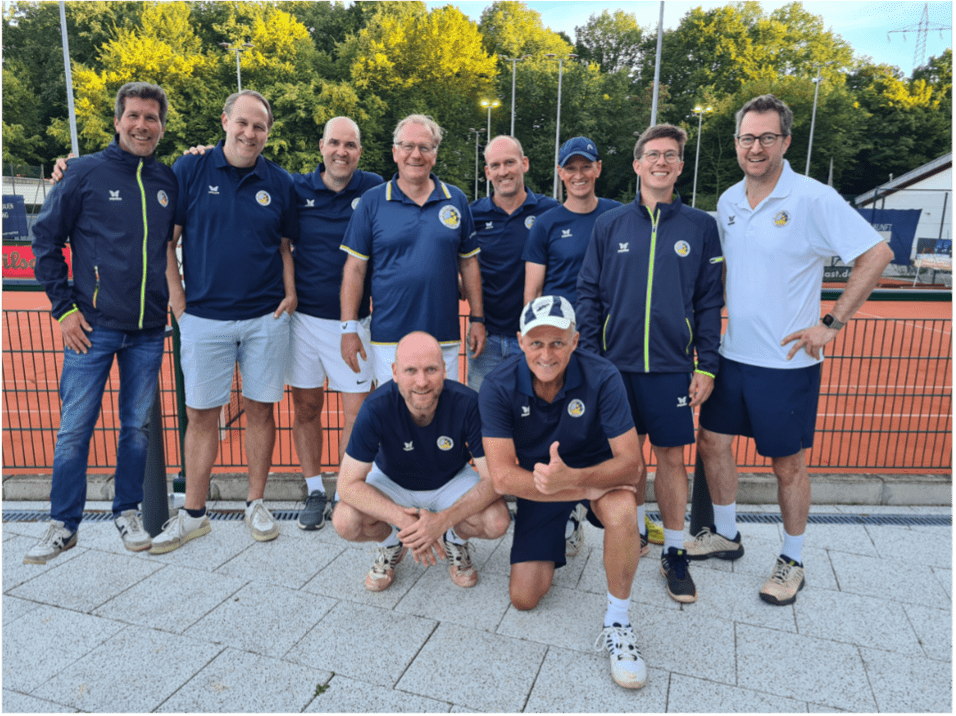 Zehn Personen in Sportbekleidung posieren l&auml;chelnd auf einem Tennisplatz mit gr&uuml;nen B&auml;umen im Hintergrund.