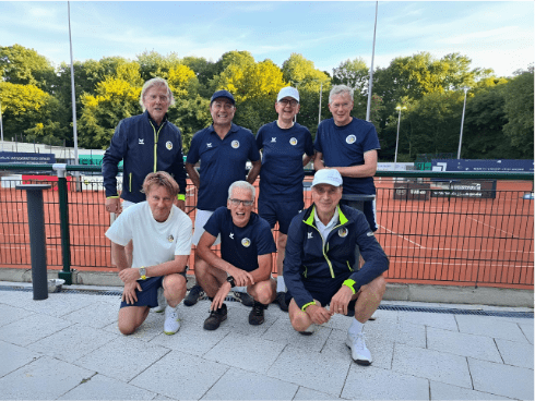 Gruppe von sechs M&auml;nnern in Sportkleidung auf Tennisplatz, posierend vor gr&uuml;ner Landschaft und roten Pl&auml;tzen.