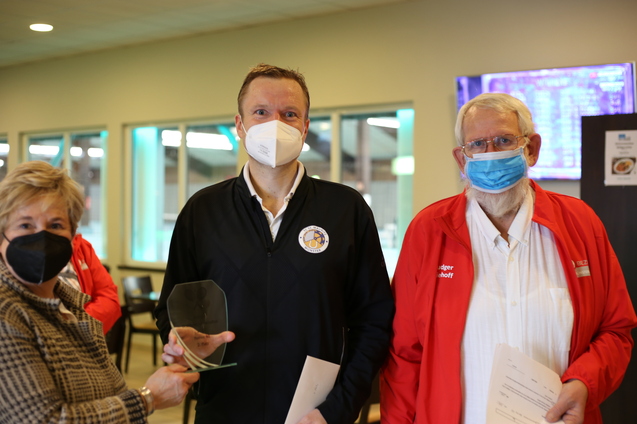 Drei Personen stehen in einem Raum, eine h&auml;lt einen Pokal, alle tragen Masken.