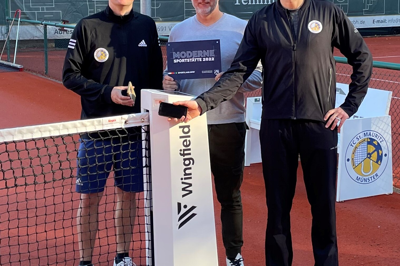 Drei M&auml;nner stehen am Netz eines Tennisplatzes, einer h&auml;lt einen Pokal, der andere ein Handy, der Dritte steht daneben.