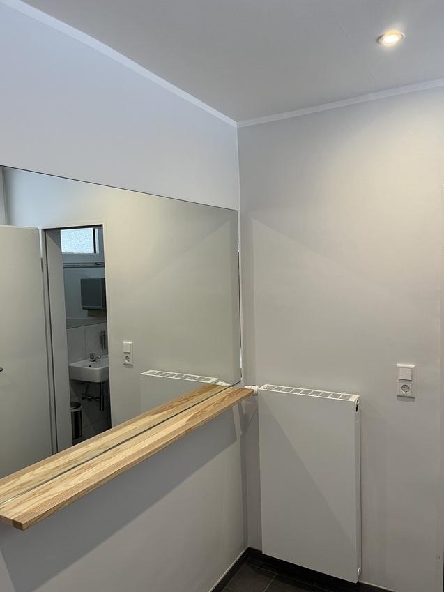 Blick in ein modernes Badezimmer mit Spiegel, Holzablage, Heizkörper und offener Tür zu einem weiteren Raum.