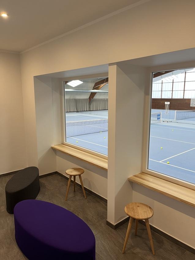 Blick auf einen Innenraum mit Holzfensterbänken und zeitgemäßen Sitzmöbeln, im Hintergrund ein Tennisplatz.