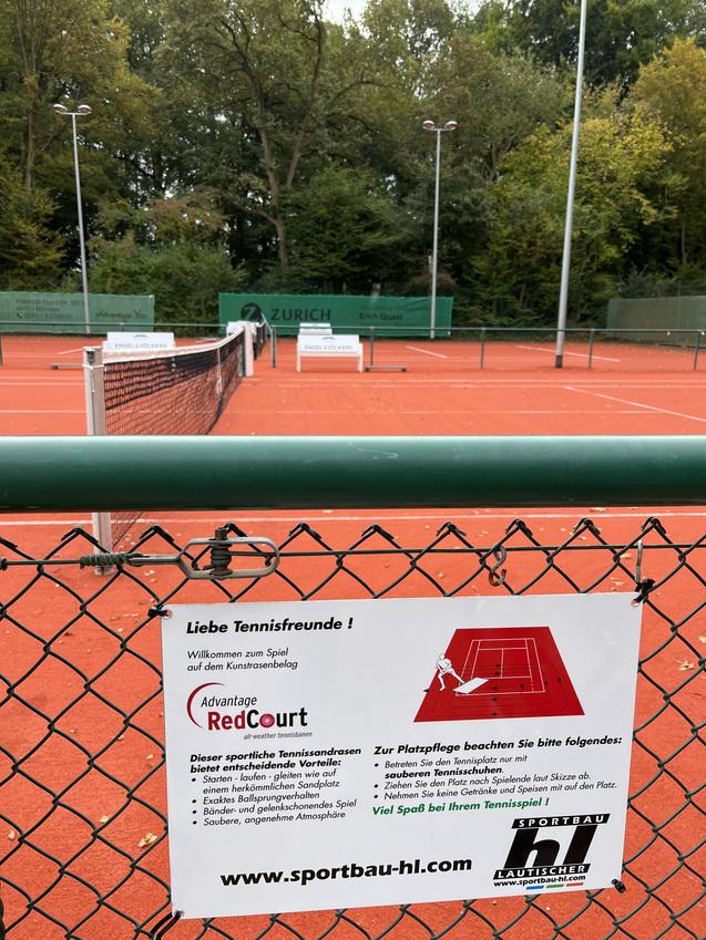 Tennisplatz mit einem Netz und Hinweisschild, umgeben von Bäumen und Sitzgelegenheiten im Hintergrund.