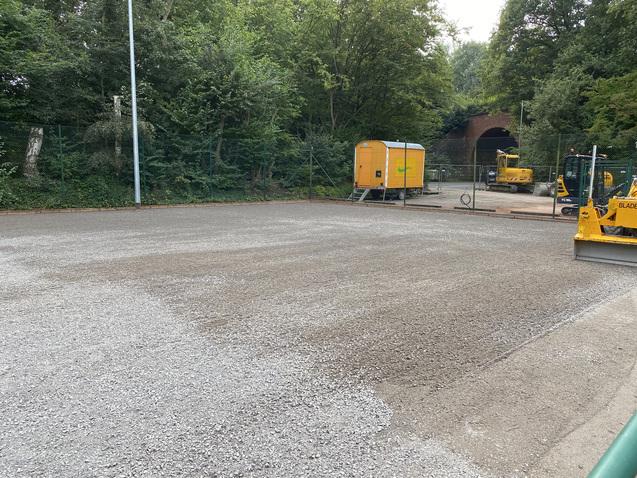 Blick auf einen geschotterten Platz, umgeben von Bäumen, mit Baugeräten und einer orangefarbenen Bauwagens im Hintergrund.