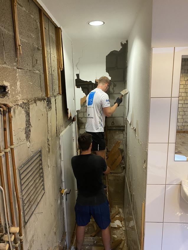 Zwei Personen renovieren einen schmalen Flur, entfernen Putz und freilegende Ziegelsteine an der Wand.