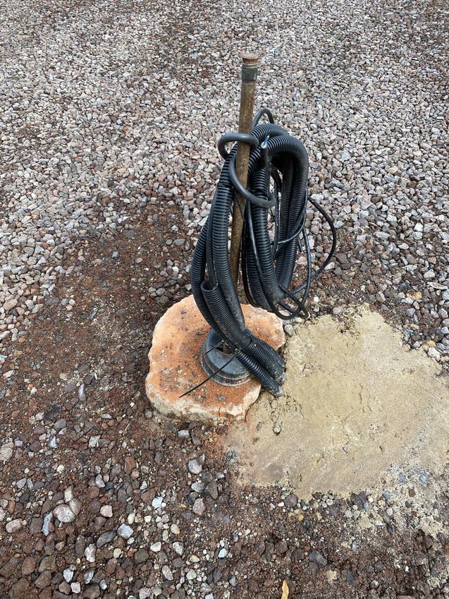 Verankerter Stab mit mehreren schwarzen Schläuchen auf einem Pflasterstein in einer kiesigen Umgebung.