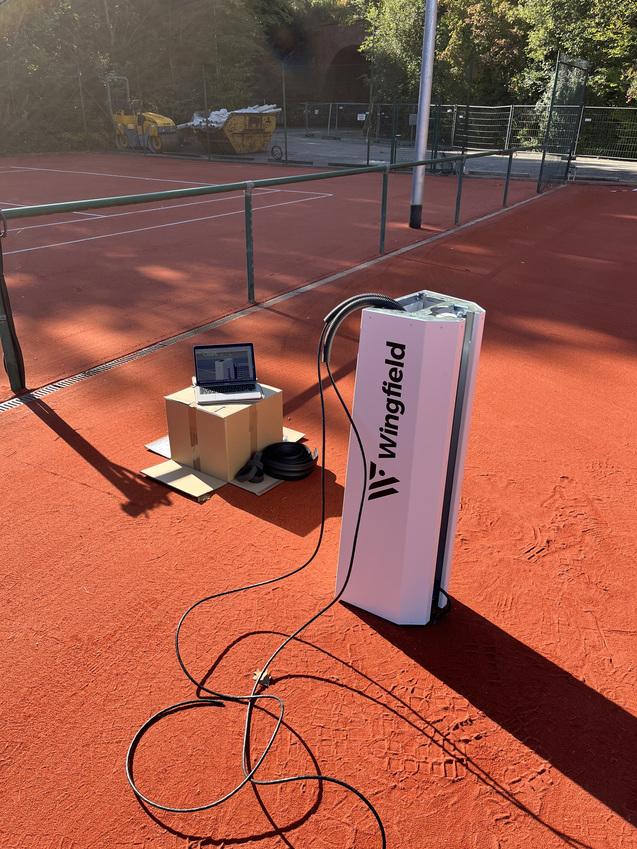 Wingfield-Anlage auf einem roten Tennisplatz mit Laptop und Kabel auf einem Karton neben der Station.