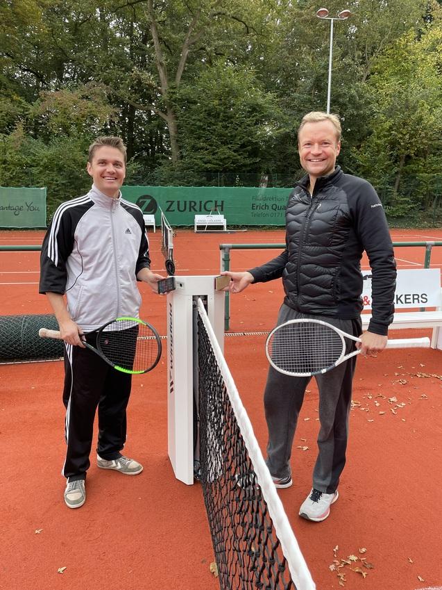 Zwei Männer mit Tennisschlägern stehen am Netz eines Sandtennisplatzes, umgeben von grünen Bäumen.