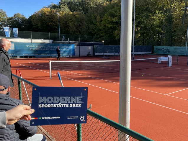Hand, die ein Schild mit der Aufschrift &bdquo;MODERNE SPORTST&Auml;TTE 2022&ldquo; h&auml;lt, im Vordergrund eines leeren Tennisplatzes.