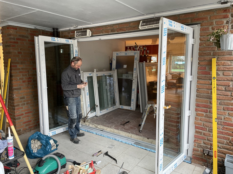 Handwerker steht vor offenen Fensterrahmen, w&auml;hrend er mit Handy arbeitet; Umgebung zeigt Baustelle mit Werkzeugen.