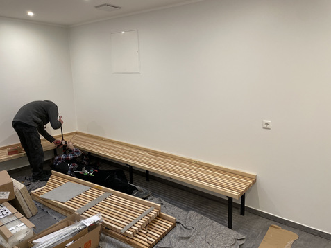 Ein Handwerker montiert eine Holzbank an einer wei&szlig;en Wand in einem Raum mit grauem Bodenbelag und Baustellenmaterial.