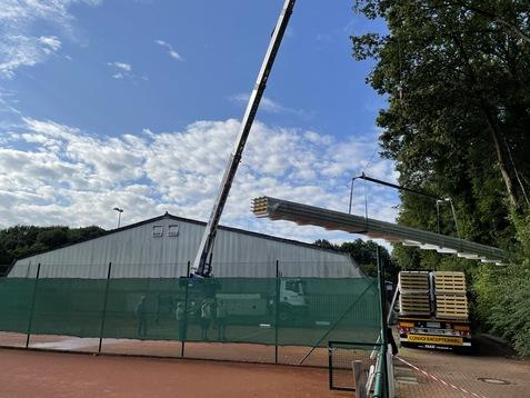 Kran hebt Metallträger über einen Tennisplatz, im Hintergrund eine Sporthalle und bewölkter Himmel.