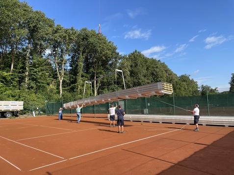 Arbeiter heben eine lange Aluminiumstruktur auf einem Tennisplatz mit rotem Belag unter blauem Himmel.