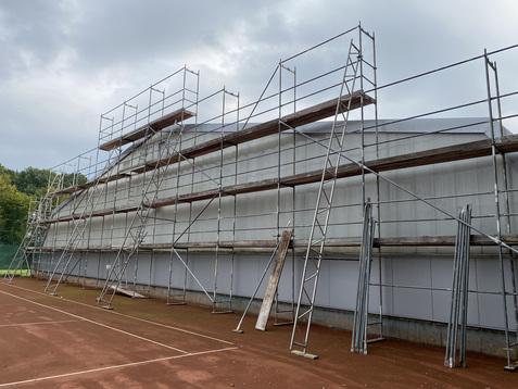 Blick auf ein gerüstetes Gebäude neben einem Tennisplatz mit rotem Sandboden. Wolken am Himmel.