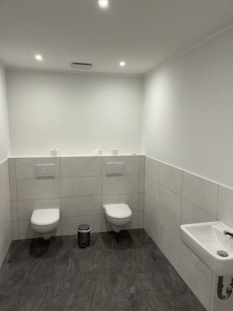 Moderne Toilette mit zwei wandhängenden Toiletten, einem Waschbecken und einem Abfalleimer auf gefliester Fläche.