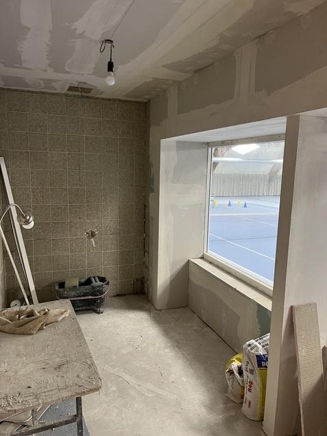 Blick auf einen unvollendeten Raum mit Fenster, das einen Tennisplatz mit Markierungen zeigt. Baustellenmaterialien sind sichtbar.