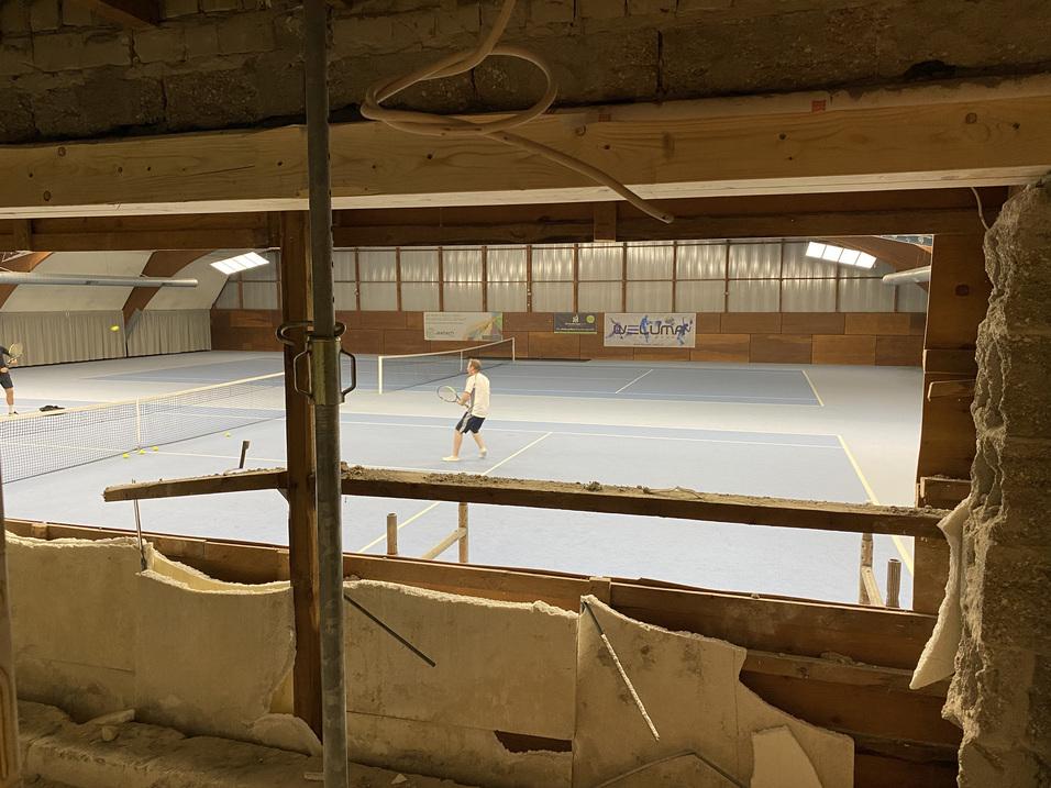 Blick durch einen offenen Wandabschnitt auf einen Tennisspieler in einer Halle mit blauer Spielfläche.
