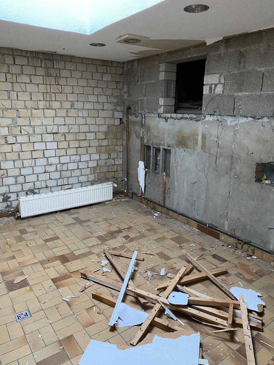 Leere, unrenovierte Zimmer mit beschädigten Wänden, einem Heizkörper und herumliegendem Baumaterial auf dem Boden.