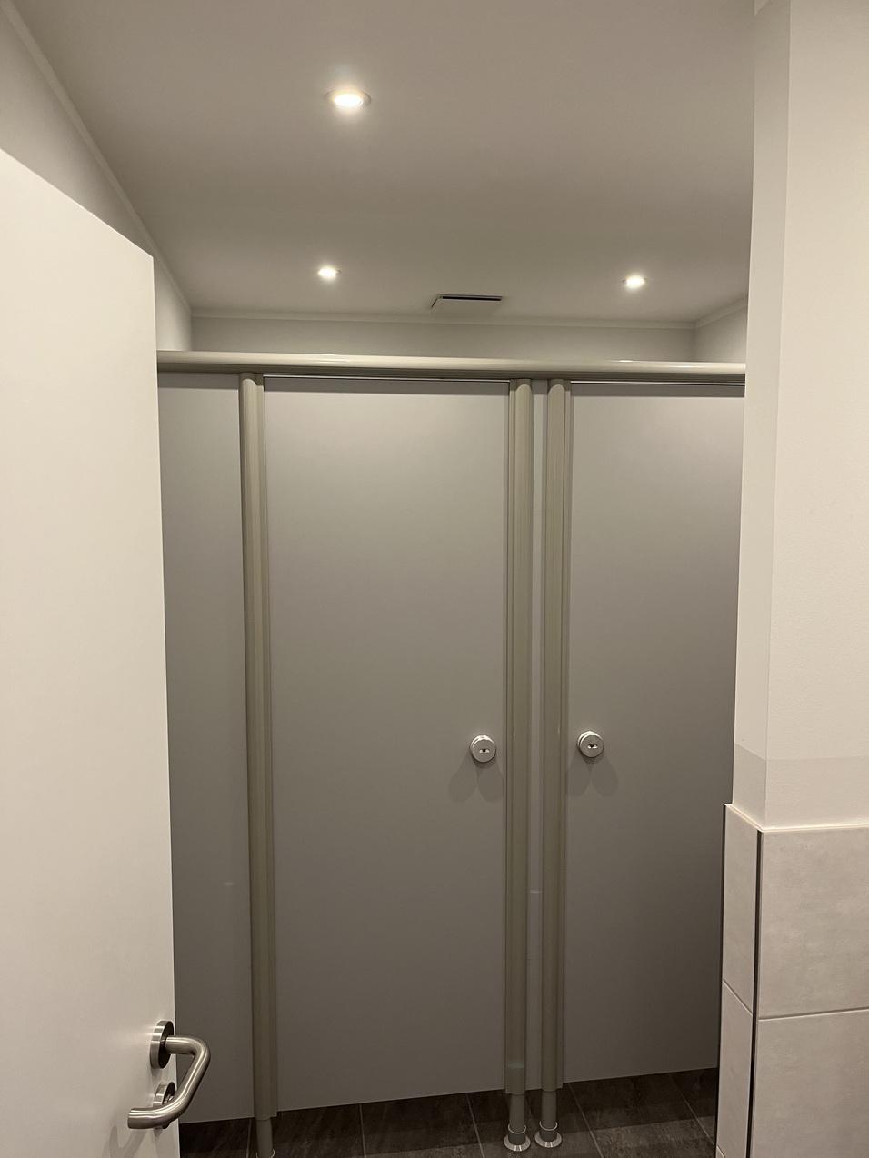 Zwei graue Trennwände von Toilettenkabinen in einem hell erleuchteten Raum.