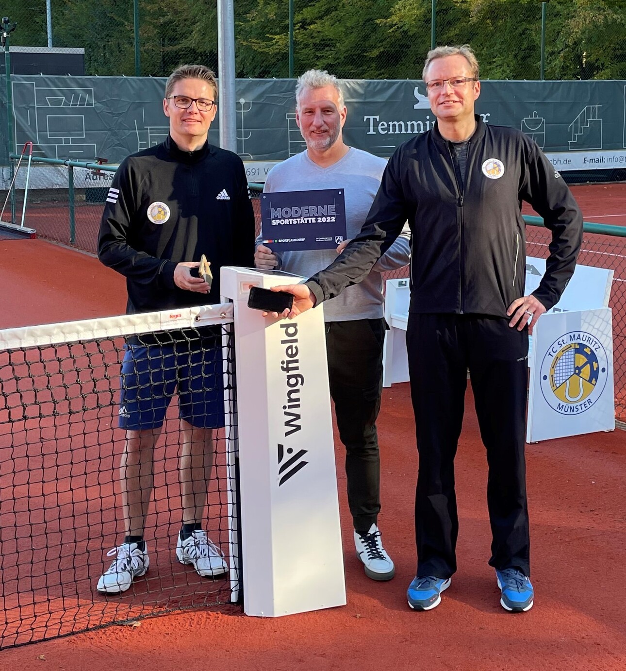 Drei M&auml;nner stehen am Netz eines Tennisplatzes, einer h&auml;lt einen Pokal, der andere ein Handy, der Dritte steht daneben.