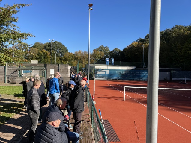 Zuschauer stehen entlang eines Tennisplatzes, umgeben von Gr&uuml;n und einer klaren blauen Himmel.